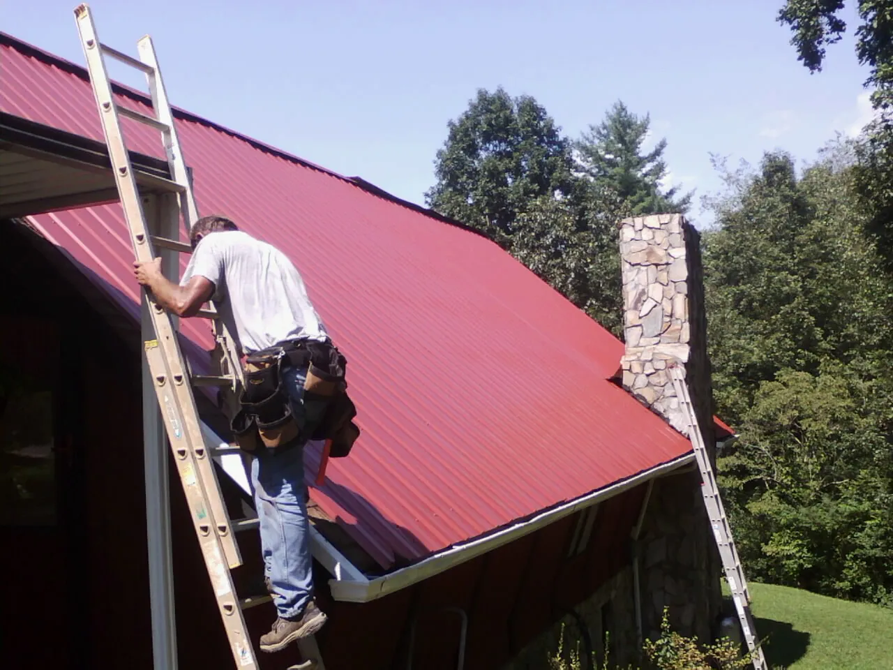 Roofer on a ladder beside a red metal roof and stone chimney — Soffit & Fascia Repair by Roofing Brothers in Hawk Springs